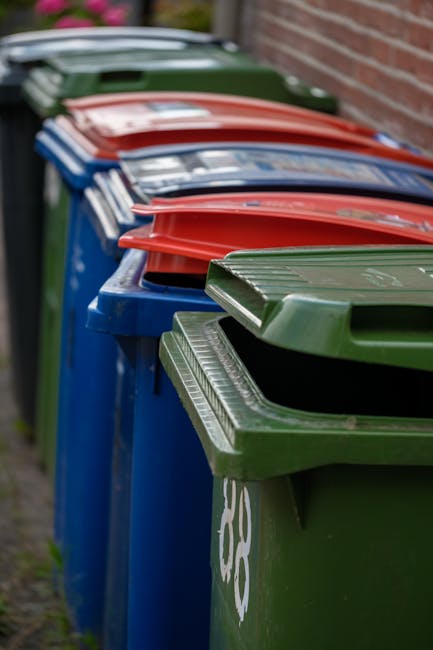 A black plastic wheelie bin with a white label and the text 'ST. JOHN'S' printed on it, positioned on a sidewalk beside a street at night. The bin's lid is open, revealing a clutter of cardboard boxes