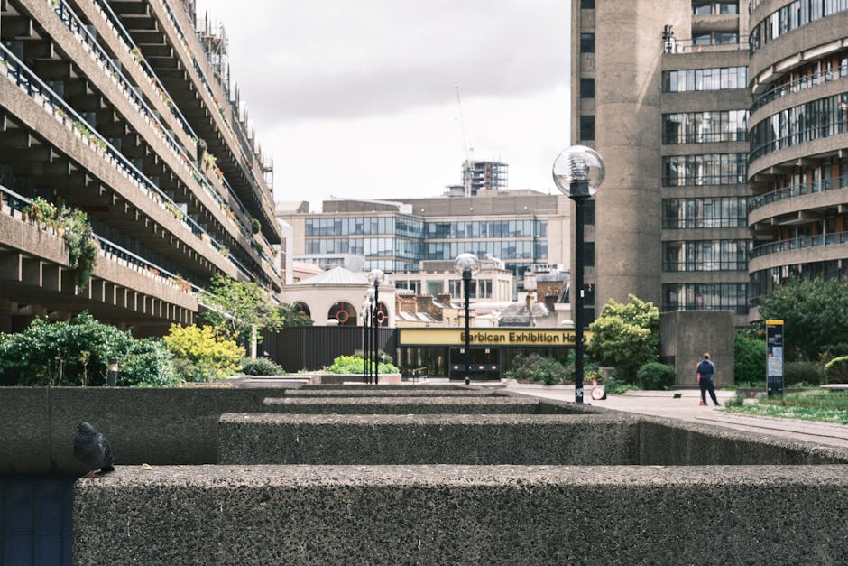 The image depicts a paved public pathway alongside a canal or river, situated in an urban setting. On the left, part of a modern building with a dark exterior and large, protruding glass windows is vi