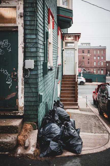 A pair of large plastic garbage bags filled with waste are positioned on the pavement next to a curb, with the bags appearing crumpled and somewhat sagging, indicating they are partially filled and re